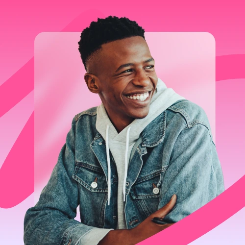 A young man with short dark hair and a wide smile looks off to his right, wearing a denim jacket over a light-colored hoodie, against a pink background.