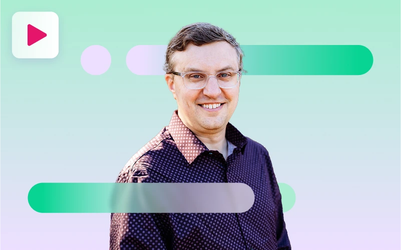 A smiling man wearing clear-rimmed glasses and a dark, patterned button-up shirt looks at the camera against a green, purple, and white abstract background with a play button icon in the top left.